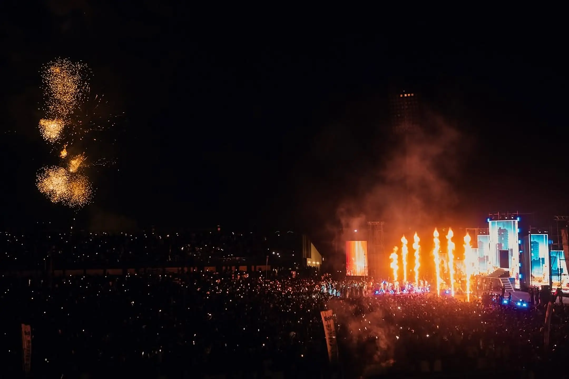 Fireworks and flame jets over a packed open-air concert at night