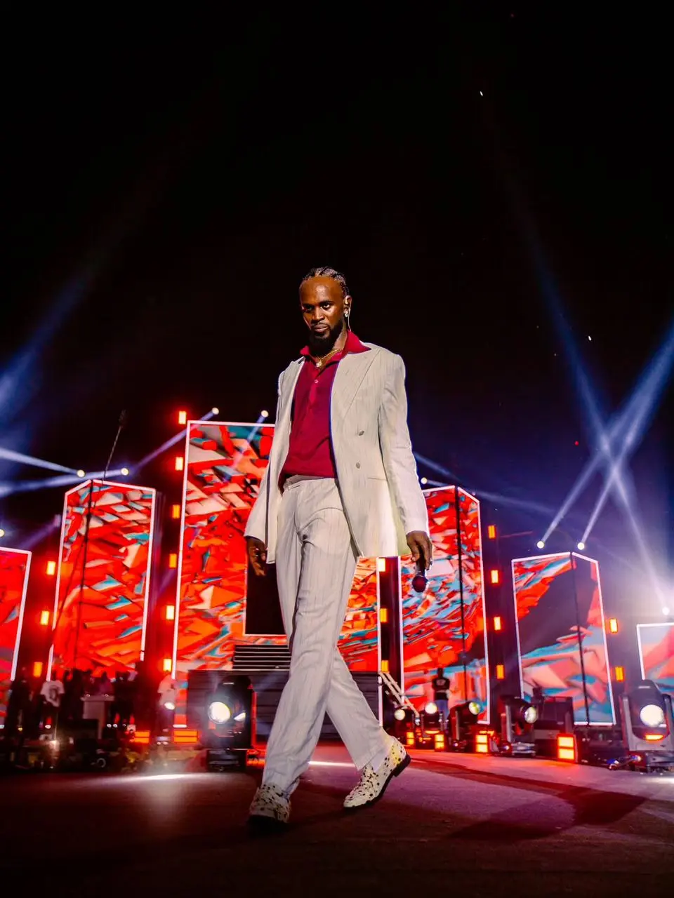 Performer in a white suit walking the stage in front of red LED screens and light beams